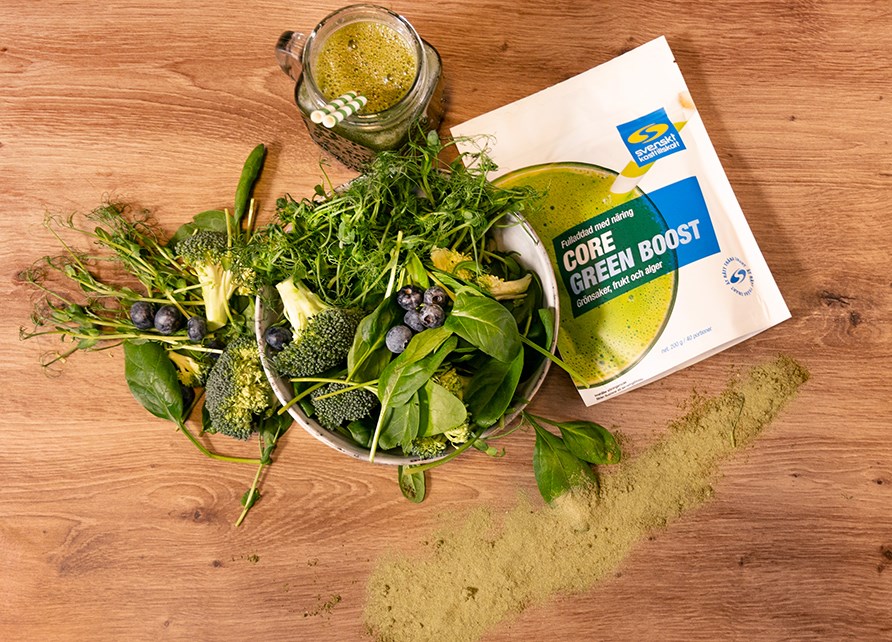 Bord med grnne bladgrntsager, en pose Core Green Boost og et glas fyldt med en grn smoothie.